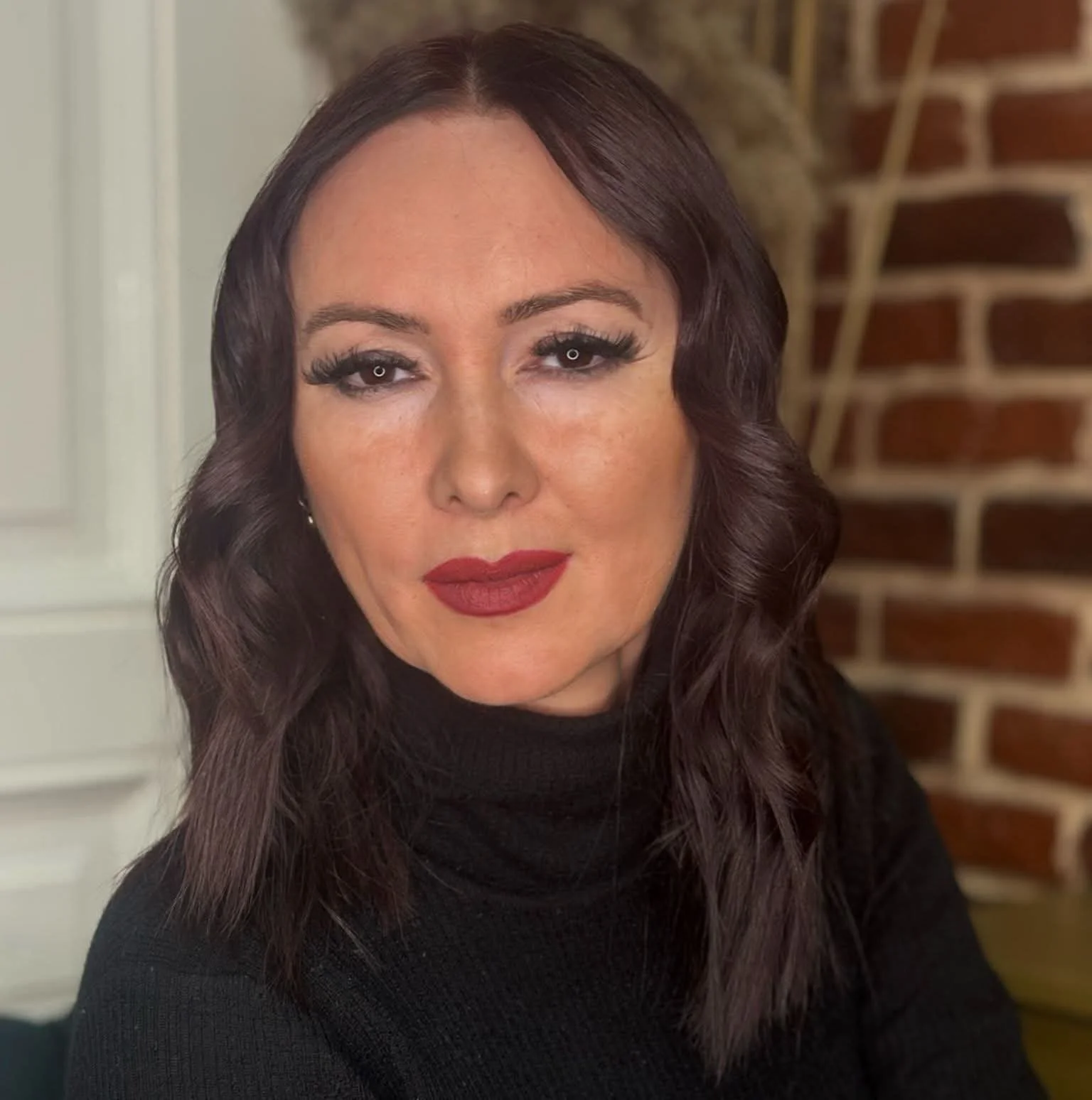Ritratto di una donna con capelli castani ondulati, trucco elegante, indossando un maglione nero, sfondo con parete di mattoni.