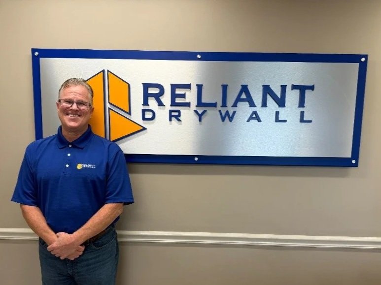 A man standing in front of a sign that reads "Reliant Drywall" with a logo of three yellow triangles.