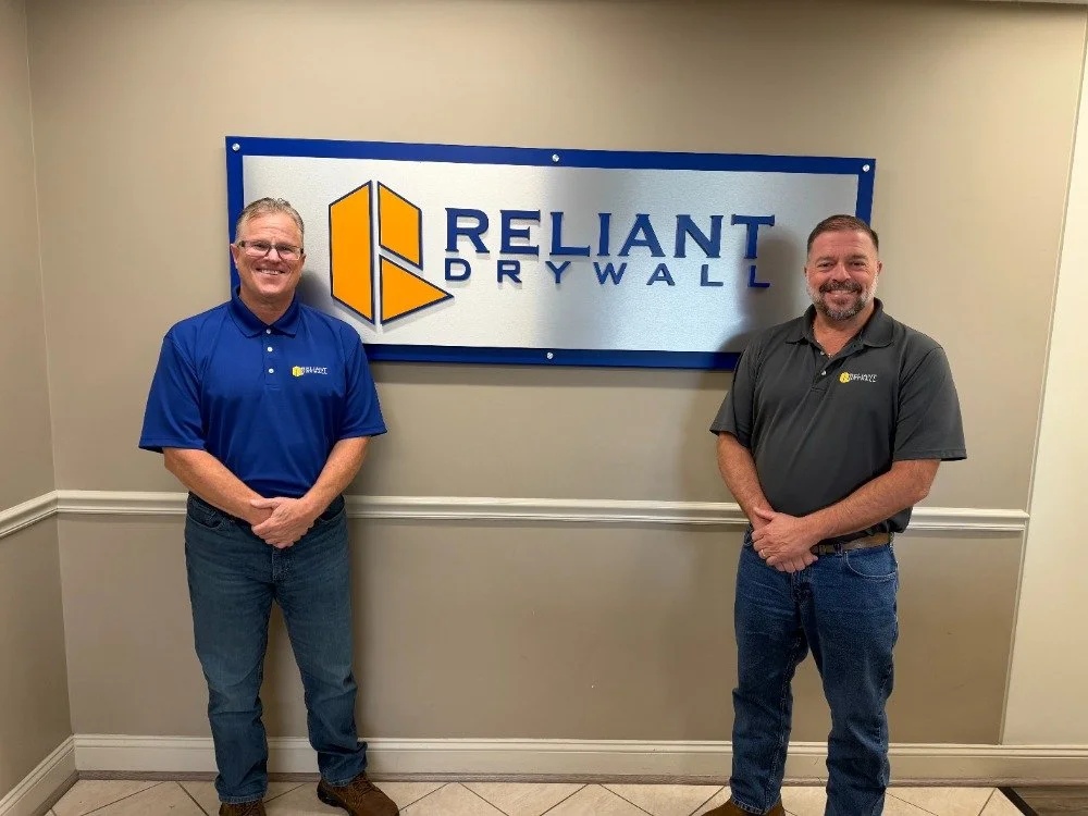 Two men standing in front of a Reliant Drywall sign, smiling, in an indoor office setting.