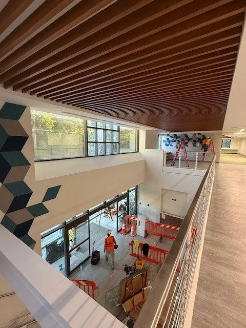 Interior view of a building under construction or renovation, showing workers on ladders, construction barriers, and tools, with large windows allowing natural light.