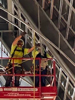 Two airport ground crew workers wearing safety vests and masks working on an airplane wing using a cherry picker lift.