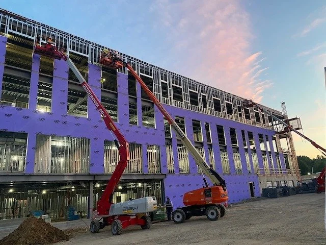 Construction site with a partially built multi-story building, purple framing, and two cherry pickers lifting workers to upper levels at dusk.