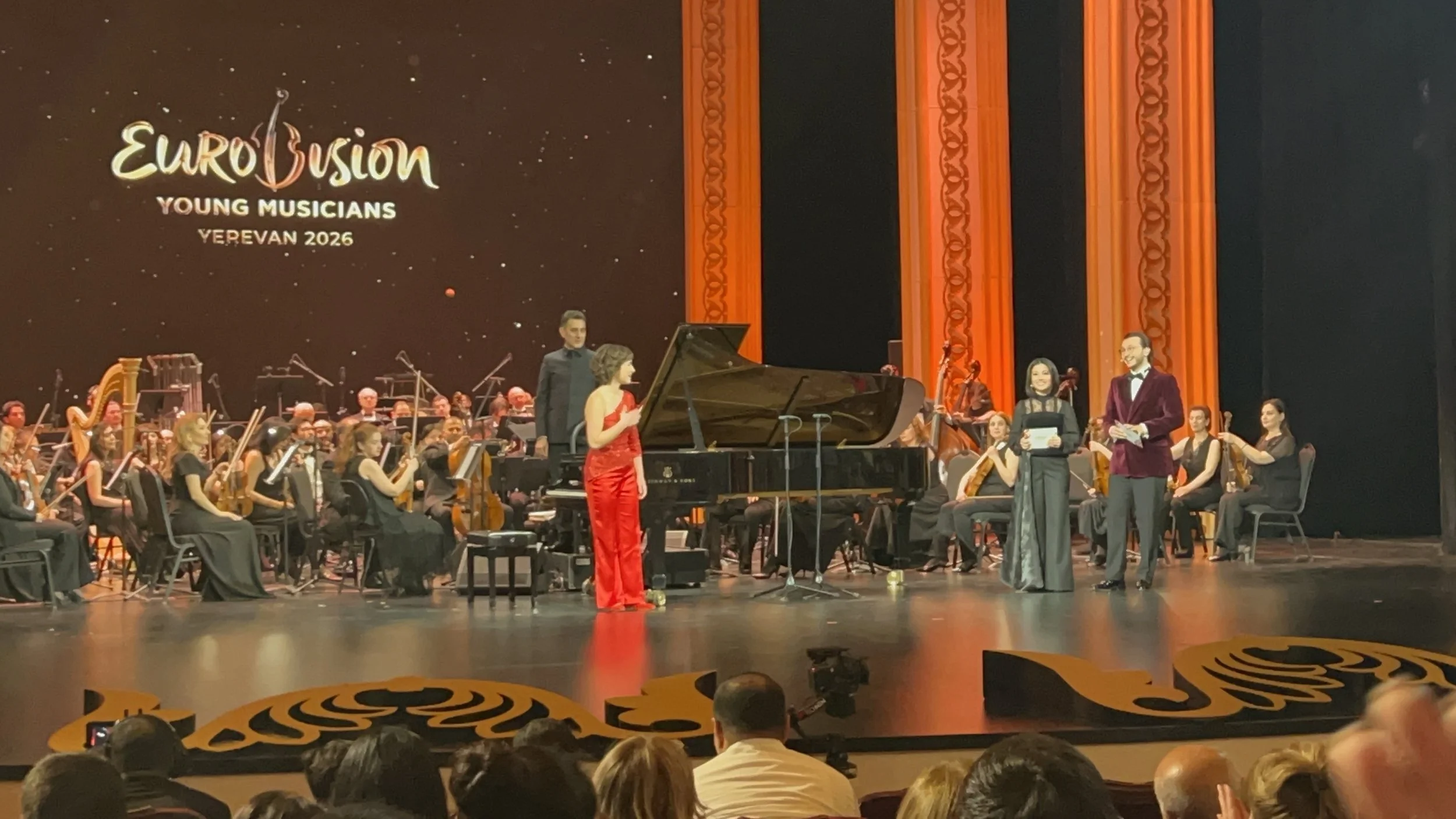 An orchestral performance at the Eurovision Young Musicians event in Yerevan 2026, featuring musicians in formal black attire, with a grand piano and a stage backdrop displaying the event logo and name.