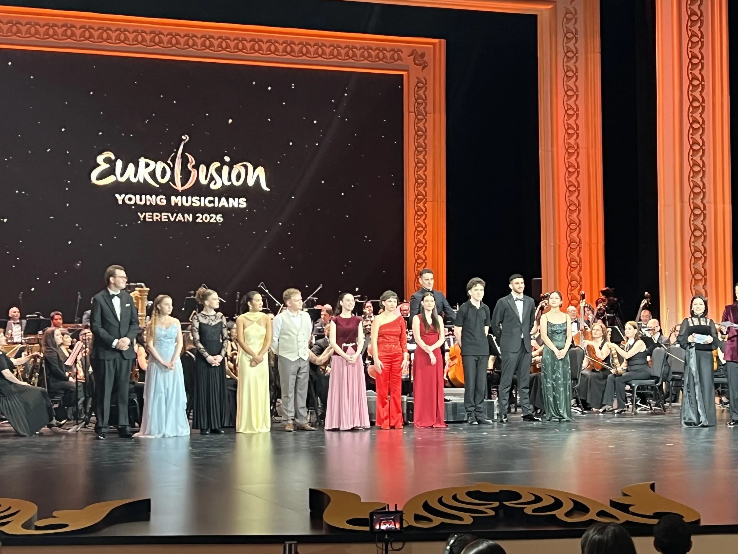 Young musicians dressed in formal attire with women in colorful evening gowns and men in black tuxedos, standing on stage at the Eurovision Young Musicians event in Yerevan 2026, with an orchestra seated behind them and a large screen displaying the 