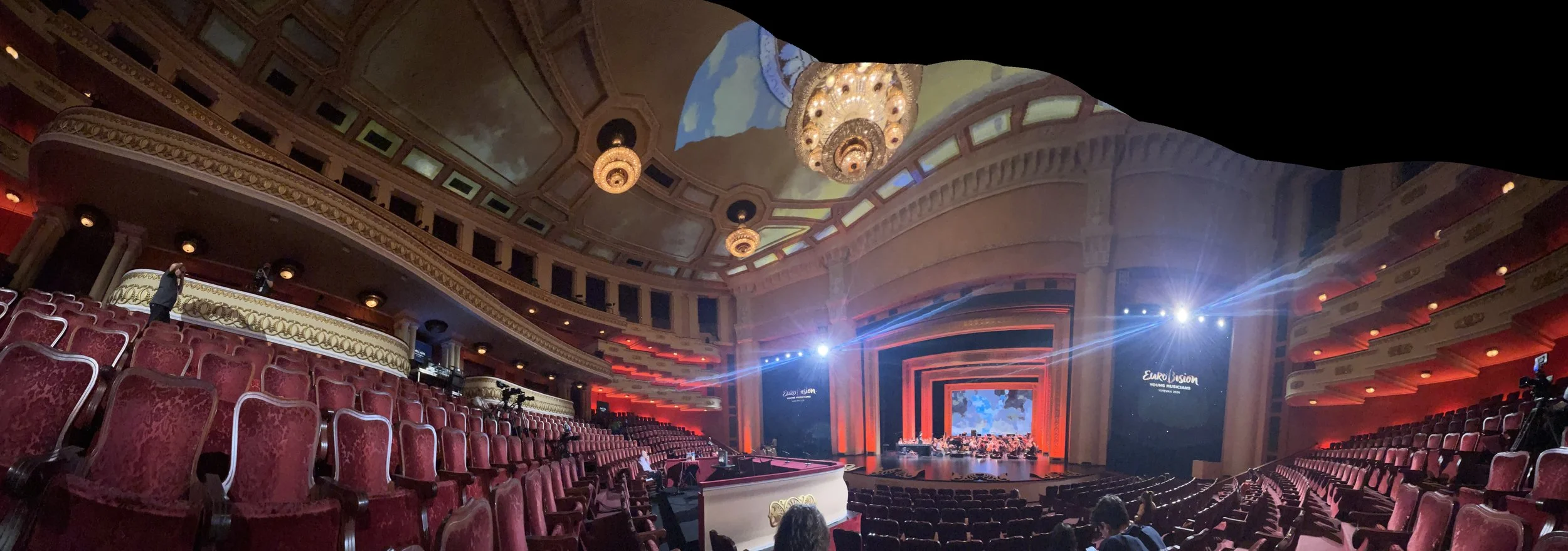 Interior of a grand theater with red velvet seats, ornate balconies, and a stage with bright lighting and a large screen displaying a colorful image. There are a few people seated and some standing, preparing for a performance.