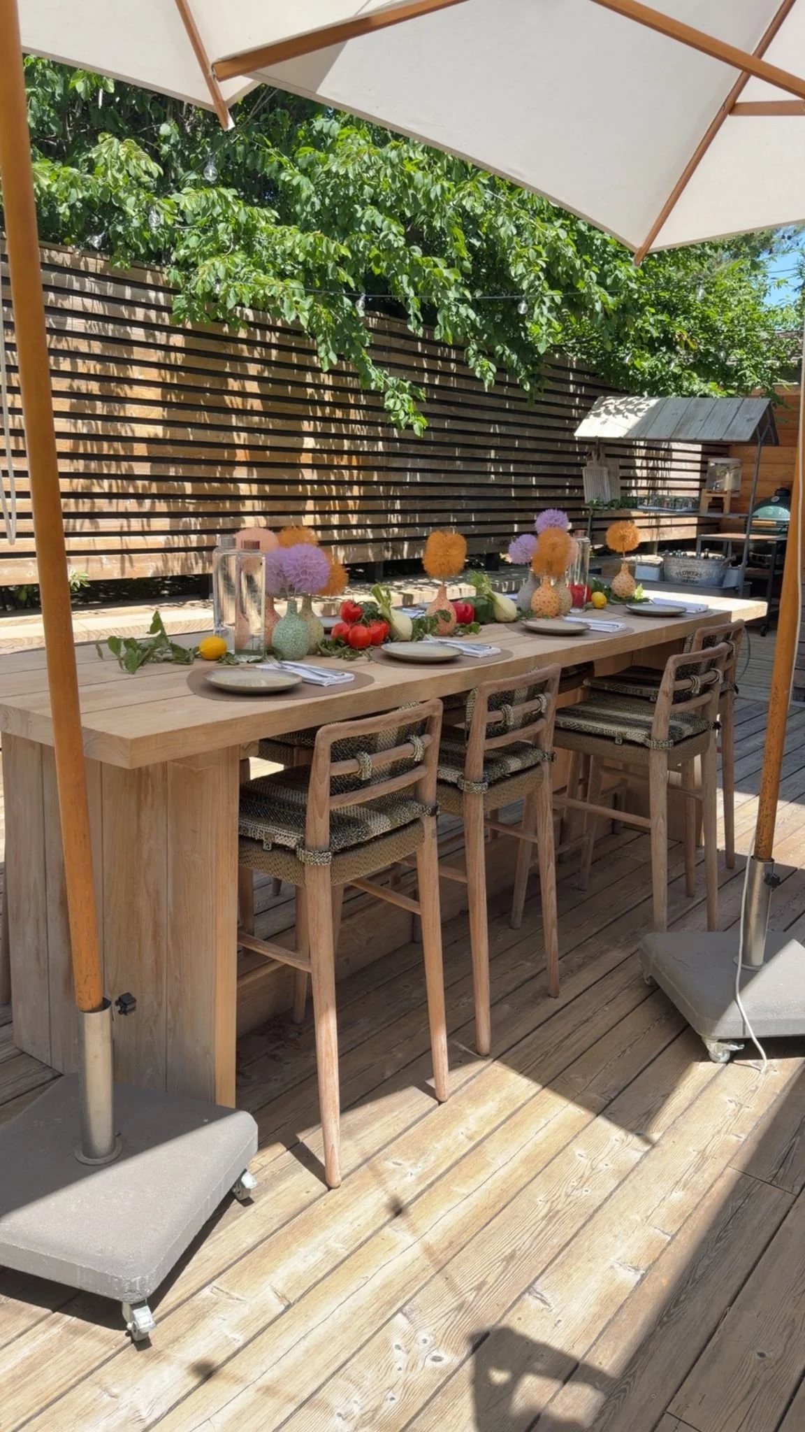An outdoor wooden dining table under a large white patio umbrella, decorated with vases of pastel-colored flowers and surrounded by wooden chairs on a wooden deck, with a wooden fence and greenery in the background.