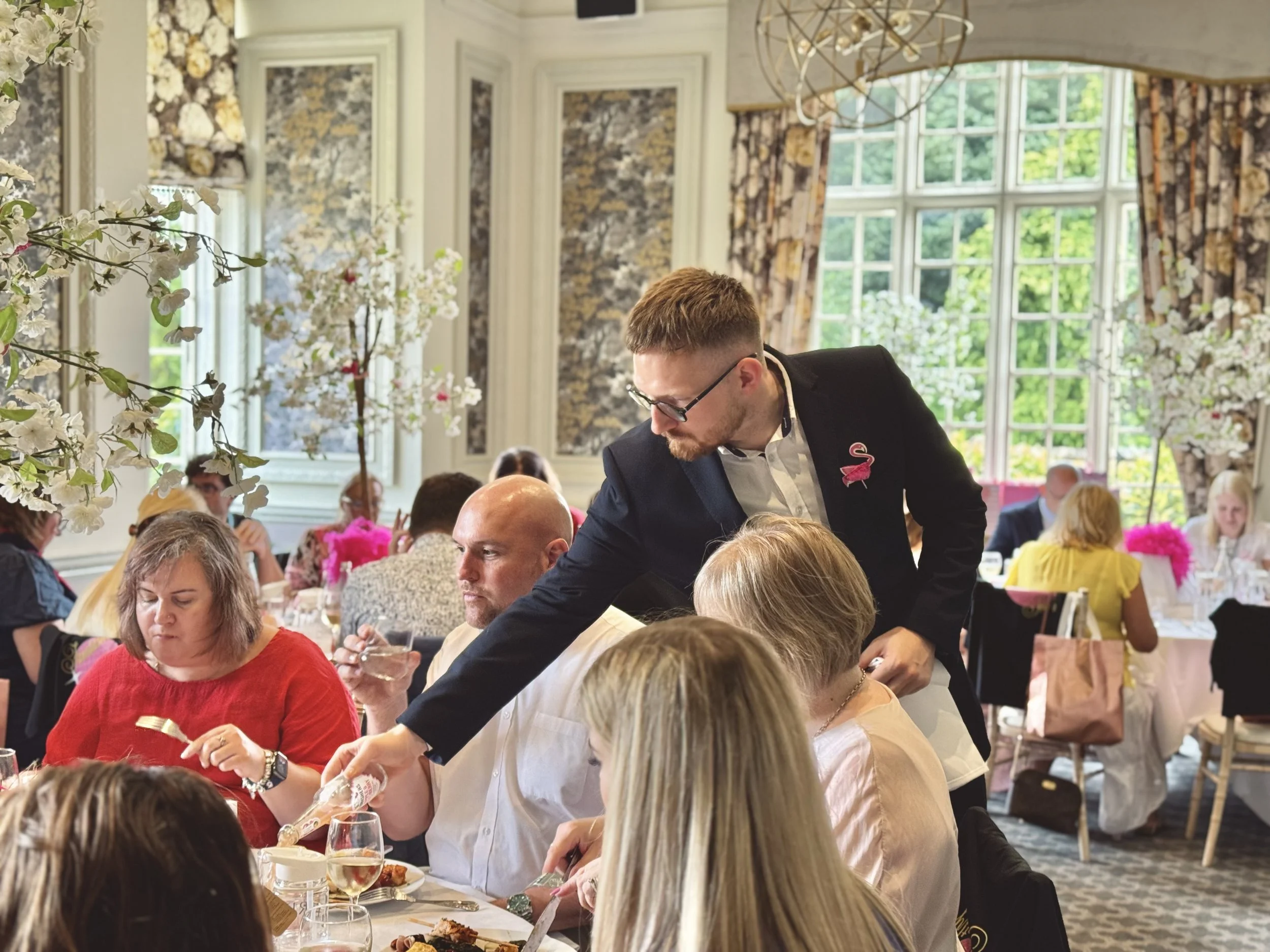 A man in a suit and glasses serving food or drinks to people at a banquet table during a formal event in a brightly decorated room with large windows and floral curtains.