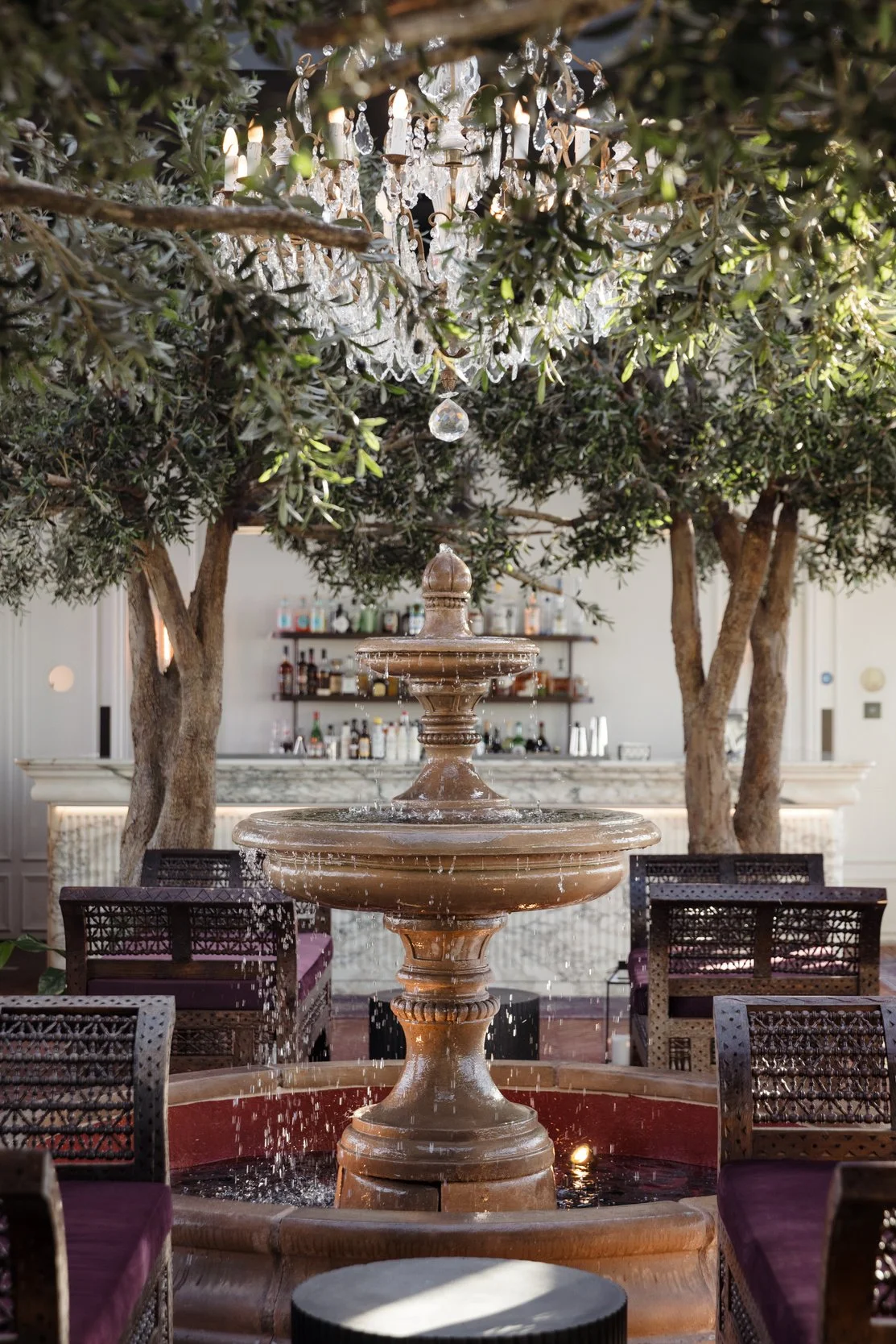Indoor scene of a tiered fountain with water cascading over it, surrounded by chairs with purple cushions, under a chandelier hanging from tree branches, with a bar in the background.