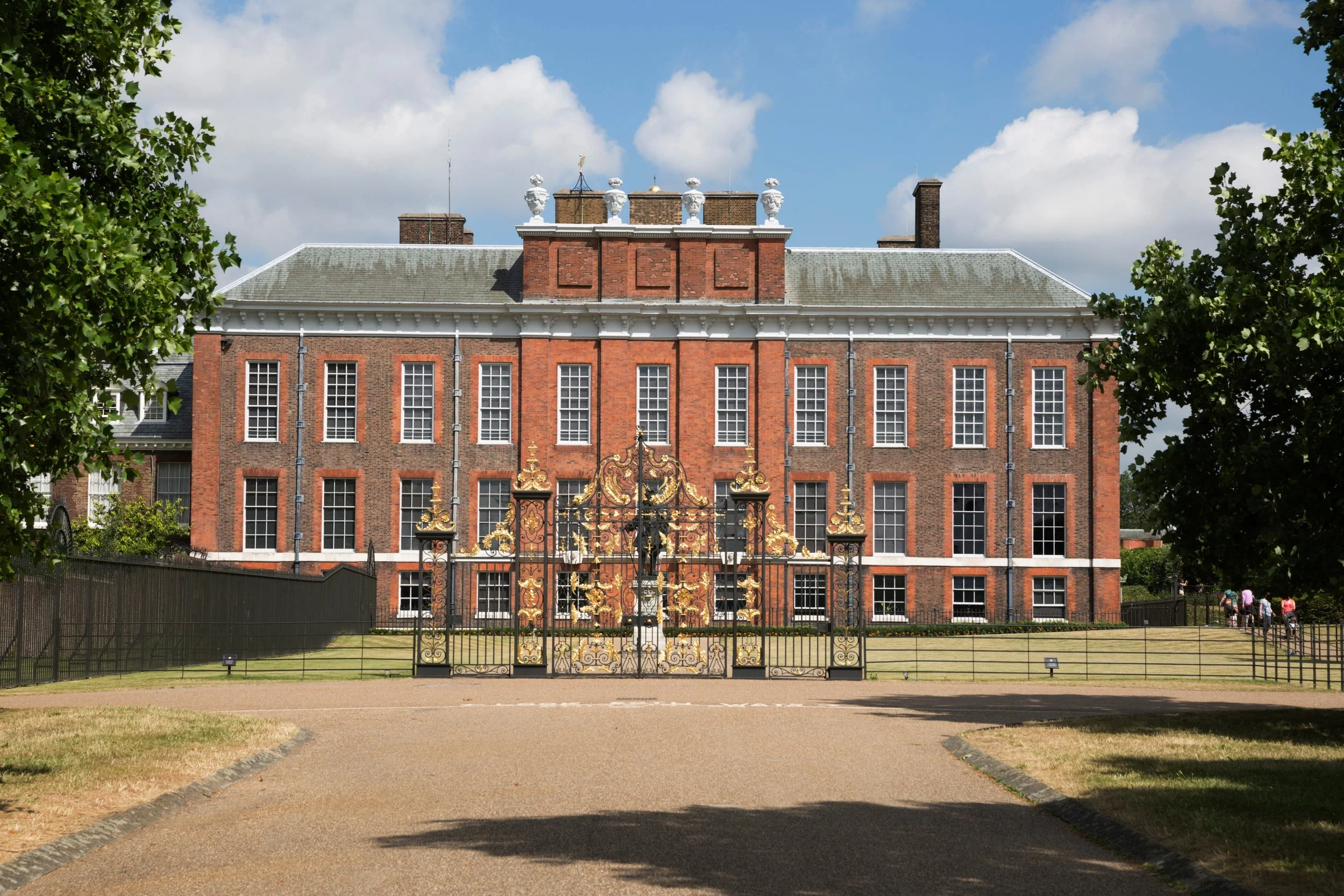 A historic red brick mansion with multiple windows and white trim, surrounded by green trees and a black iron gate, under a partly cloudy blue sky.