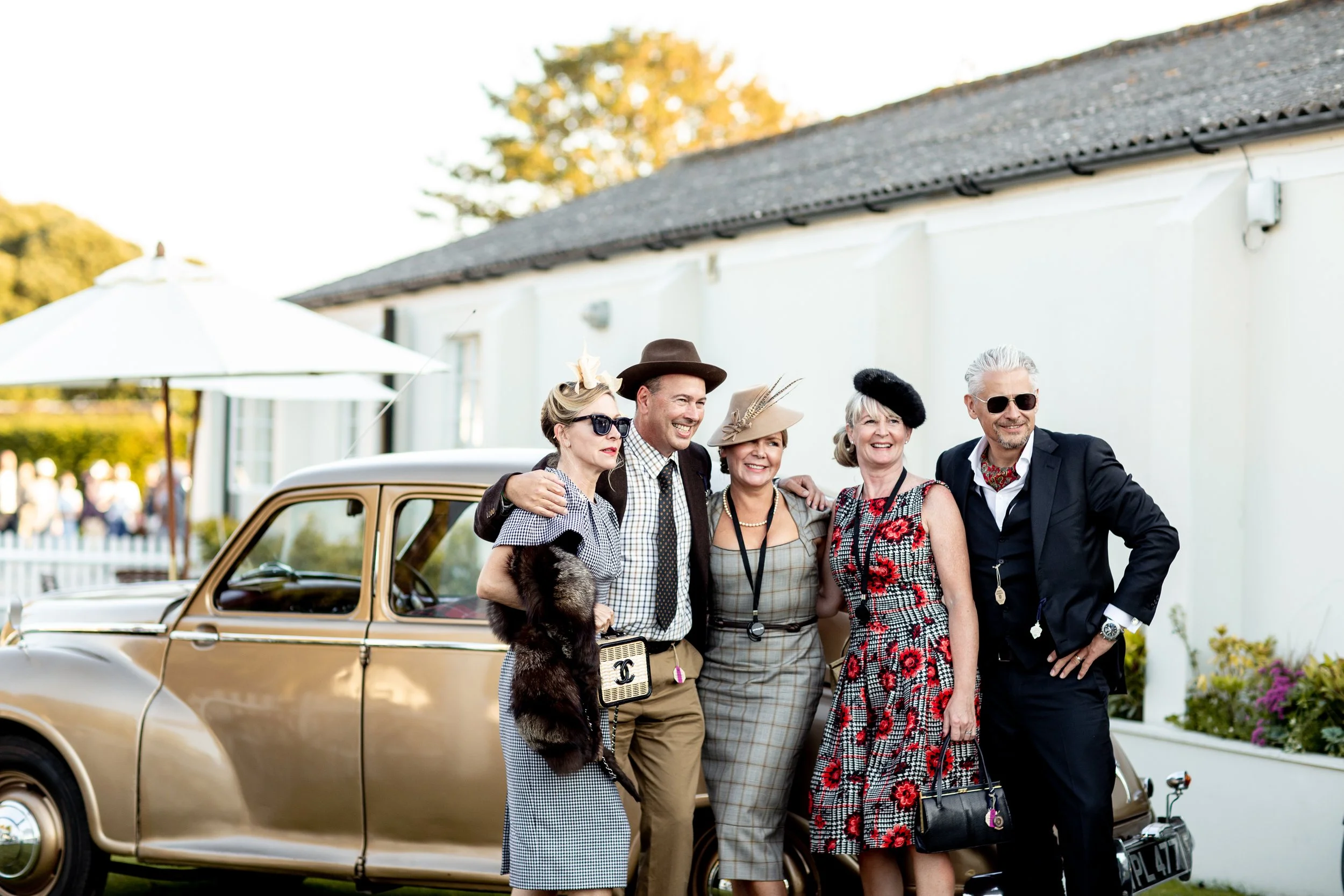 Group of five elegant and stylishly dressed older adults standing outside next to a vintage beige car, posing for a photo at a social event in the daytime, with a white building and umbrellas in the background.