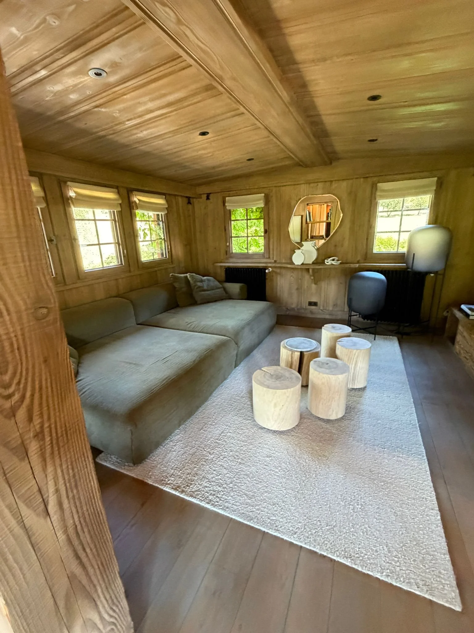 A cozy living room with wood-paneled walls and ceiling, three windows with white blinds, a large beige sectional sofa, small log-style stools, a white rug, and a small desk with a chair.