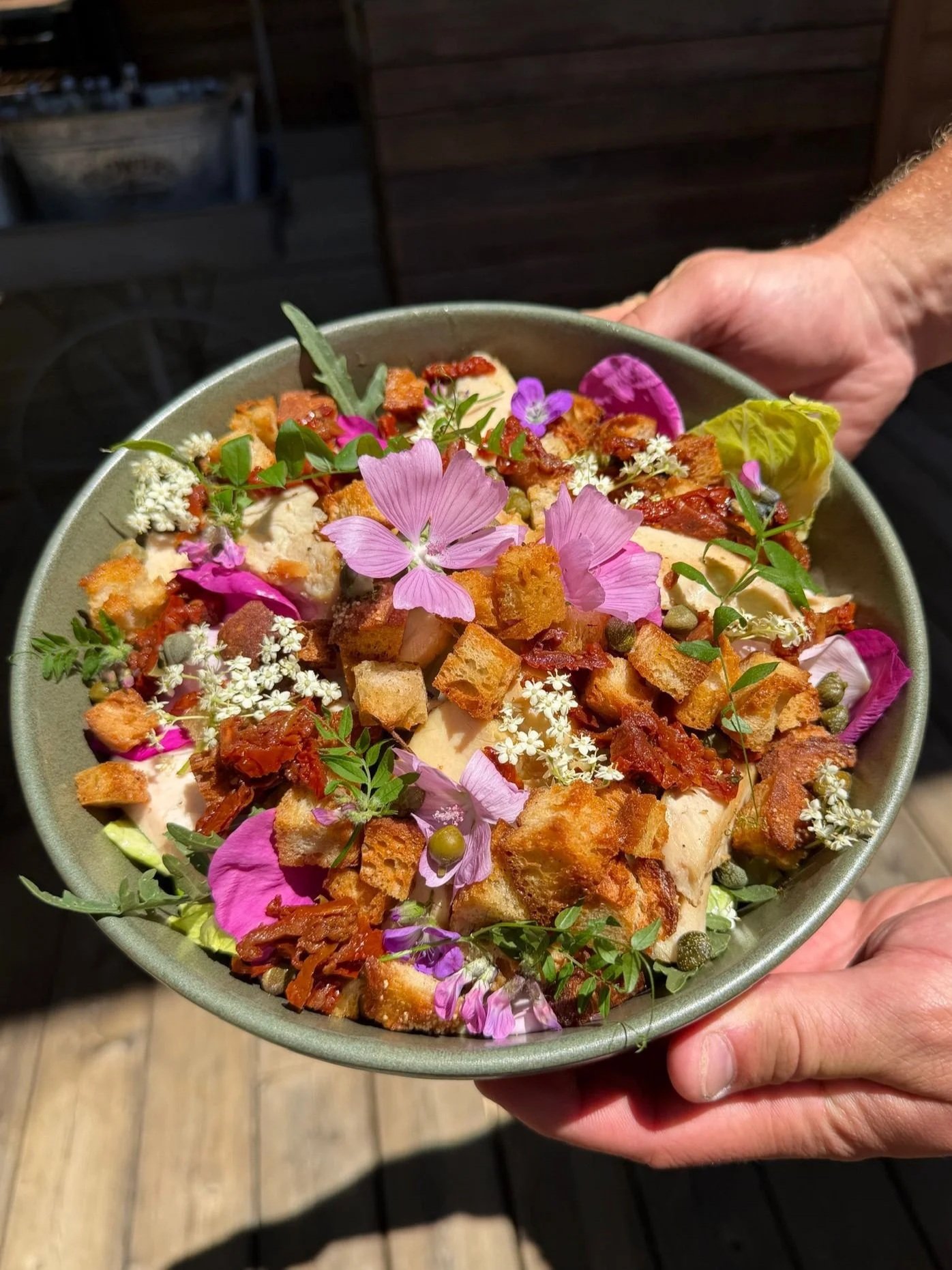 A bowl of salad topped with edible flowers, croutons, and small pieces of crispy bacon, held in a person's hand.