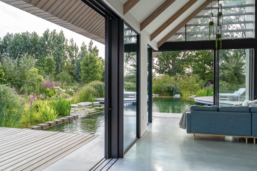 Interior view of a modern house with large glass sliding doors leading to a garden with a pond and lush greenery.