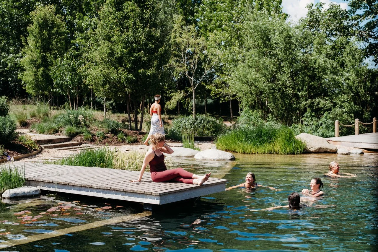 People relaxing and swimming in a peaceful outdoor pond surrounded by trees and greenery on a sunny day