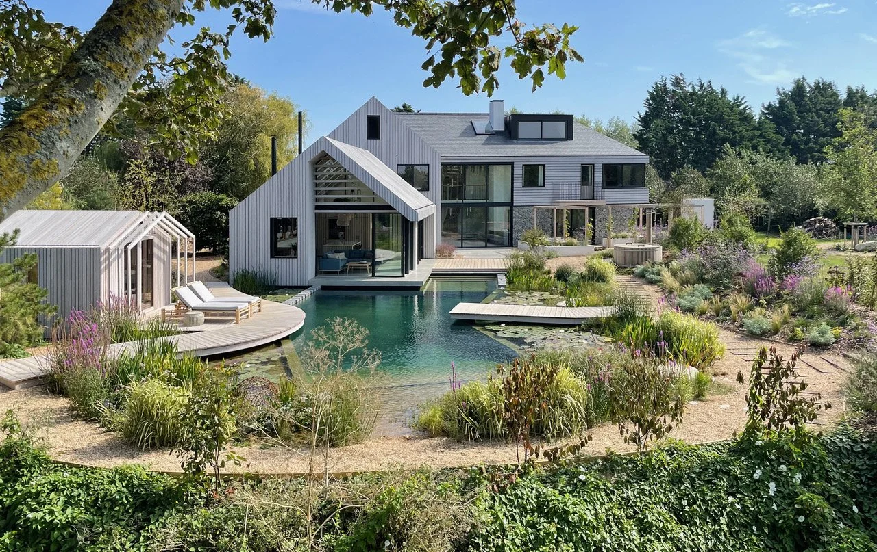 Modern house with multiple levels and large windows, surrounded by lush greenery, with a swimming pool in the foreground and a landscaped garden.