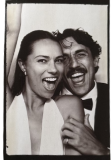 Black and white photo of a joyful woman and man smiling and laughing closely together, with the woman showing her teeth and the man with a mustache and curly hair.