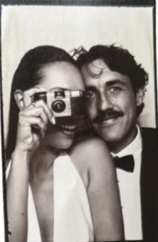 A woman and a man taking a selfie together, smiling, with the woman holding a camera and the man wearing a tuxedo.