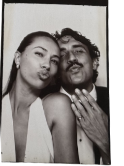 A woman and a man posing together, making kissy faces at the camera. The woman has straight hair and is wearing earrings, while the man has curly hair and is wearing a tuxedo.