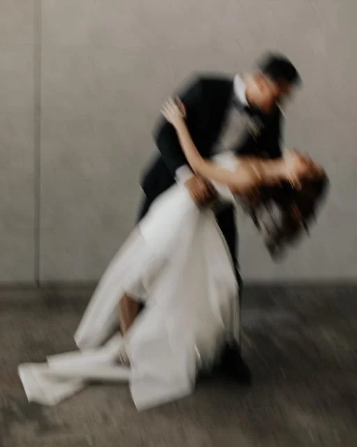 A woman in a white gown or wedding dress is leaning back and being kissed or embraced by a man in dark clothing, against a plain wall background.