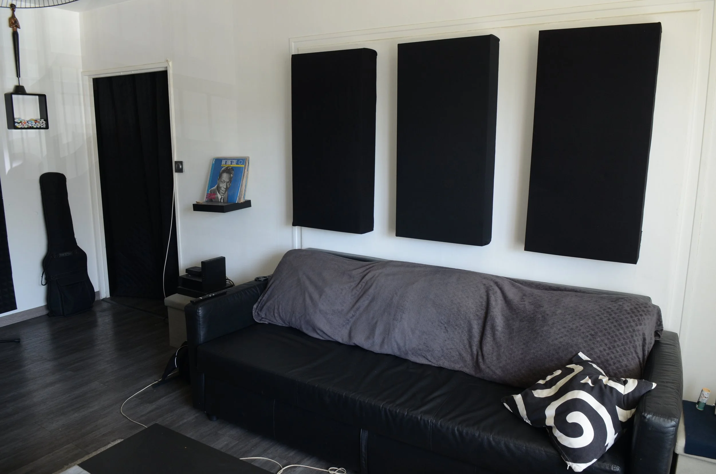 Une chambre avec un canapé, un lit avec une couverture grise, des panneaux acoustiques noirs sur le mur, une affiche de Elvis Presley, une guitare dans un étui, et des objets divers sur une table et à côté du lit.