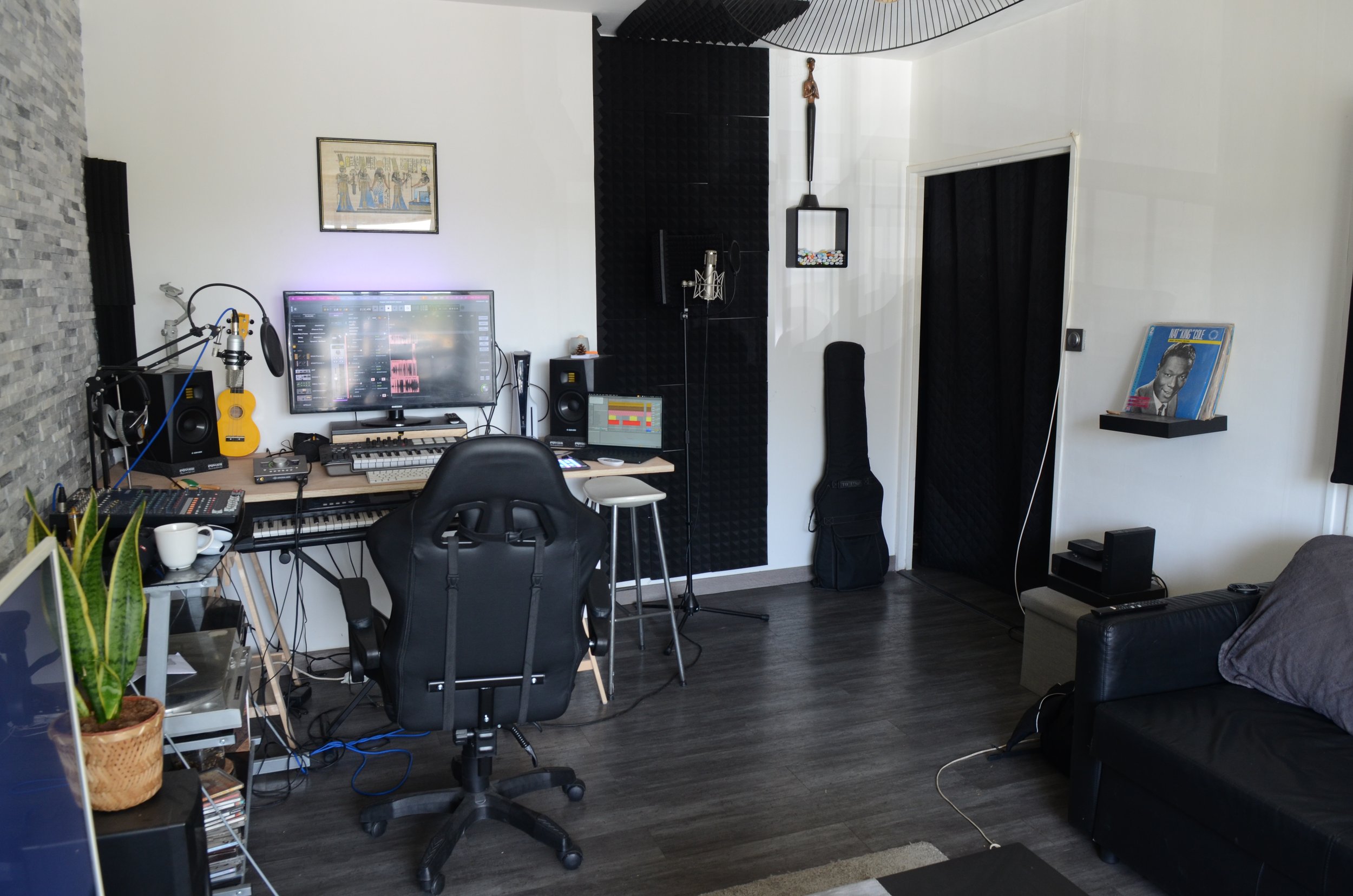 Espace de création musicale avec un bureau, un ordinateur, une tablette, un clavier, un microphone, des enceintes, une guitare acoustique, une guitare électrique, un casque audio, un instrument à cordes et des éléments de décoration, dans une pièce avec un mur en pierre et des murs blancs.