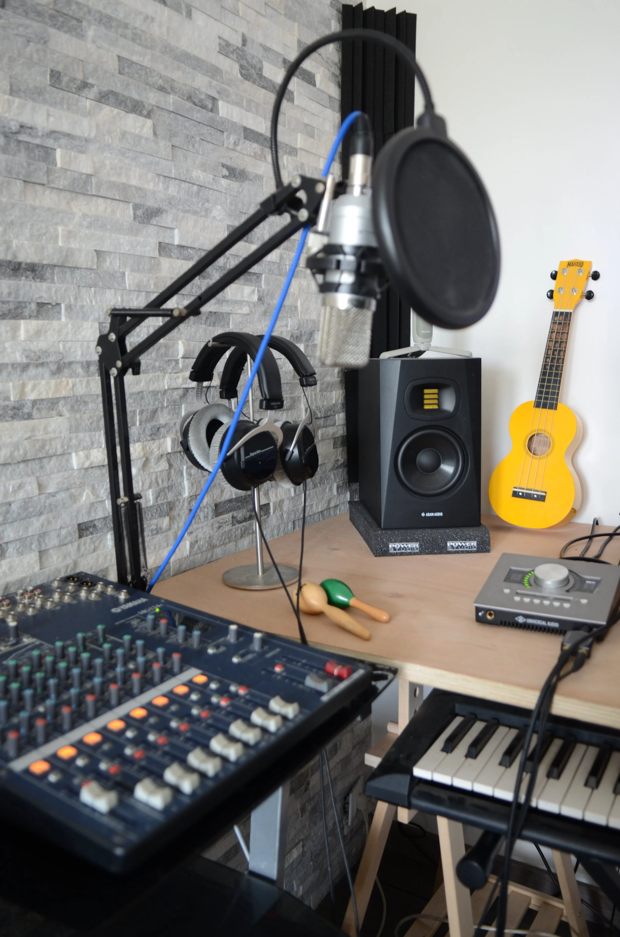Un espace de studio avec un microphone, un casque, des haut-parleurs, un clavier électronique, une console de mixage, une guitare jaune, et une arrangement d'instruments de musique sur un bureau en bois. Mur en pierre en arrière-plan.