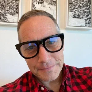 NIL expert Bill Carter wearing black glasses and a red plaid shirt, smiling slightly, with framed snowy winter landscape pictures on the wall behind him.