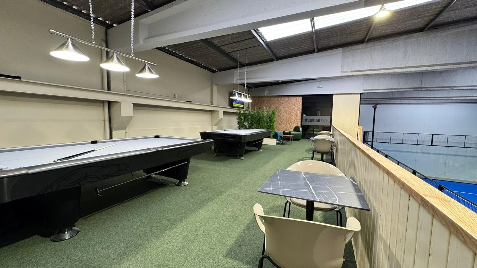 Área de lazer com mesas de sinuca e pátio interno, decorada com plantas e mobília moderna, com vista para uma quadra de tênis coberta.