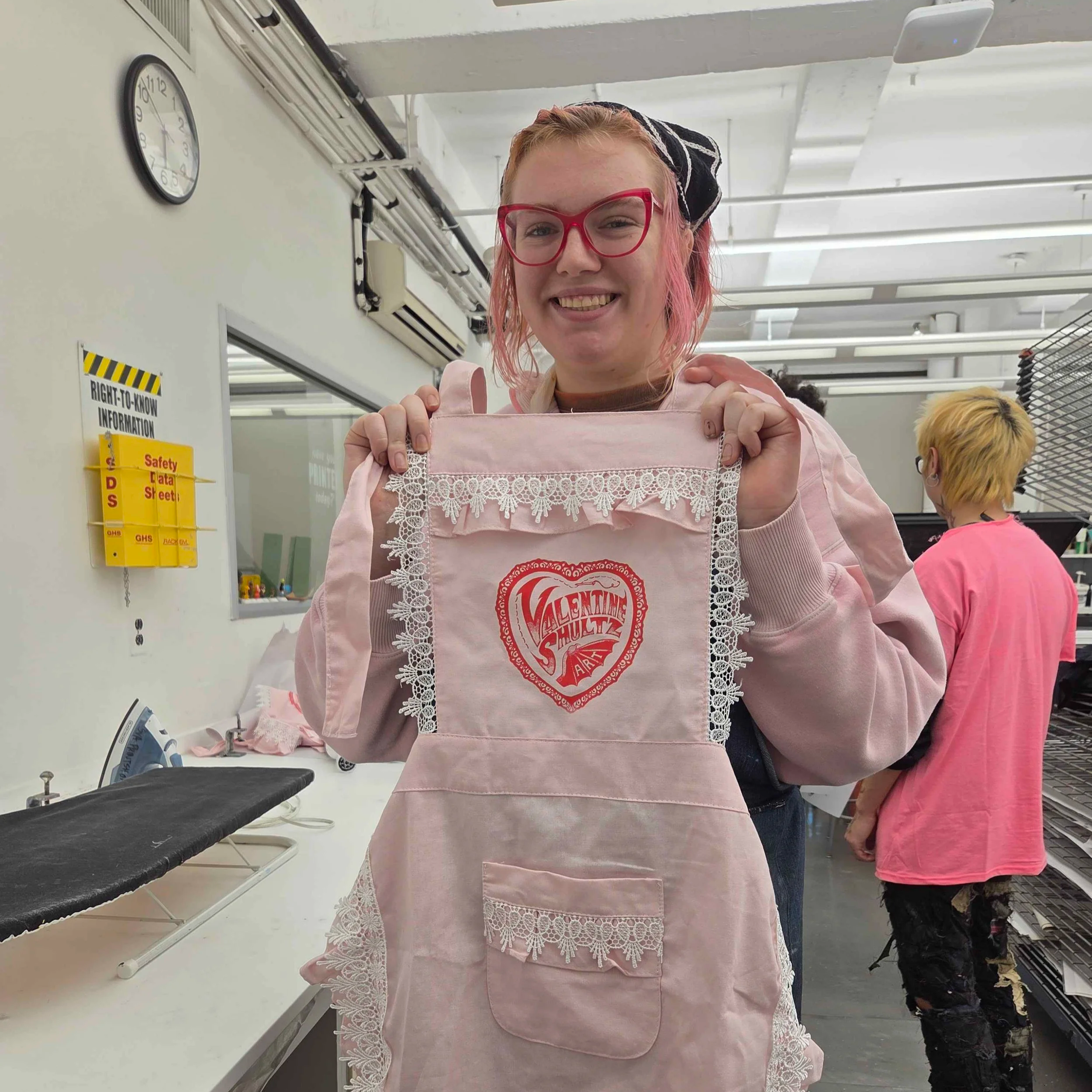 A smiling woman with pink hair and red glasses holding a pink apron with lace detailing in a store or workshop. There are other people and shelves with various items in the background.