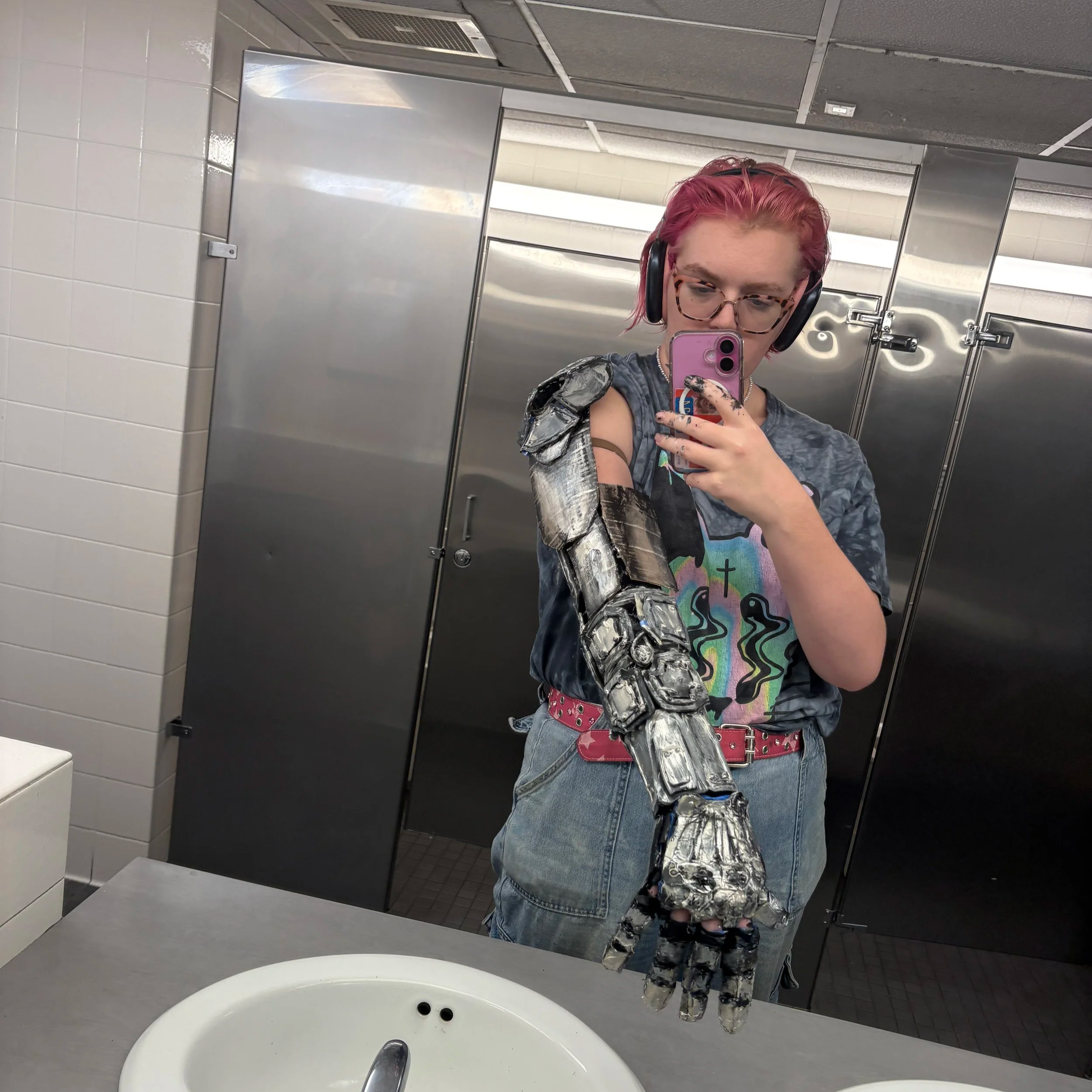 A person with pink hair, glasses, and headphones taking a selfie in a bathroom mirror. The person is wearing a colorful shirt, jeans, and a pink belt. There is a metallic prosthetic arm visible, and the bathroom has white tiled walls, a stainless steel stall, and a white sink in the foreground.