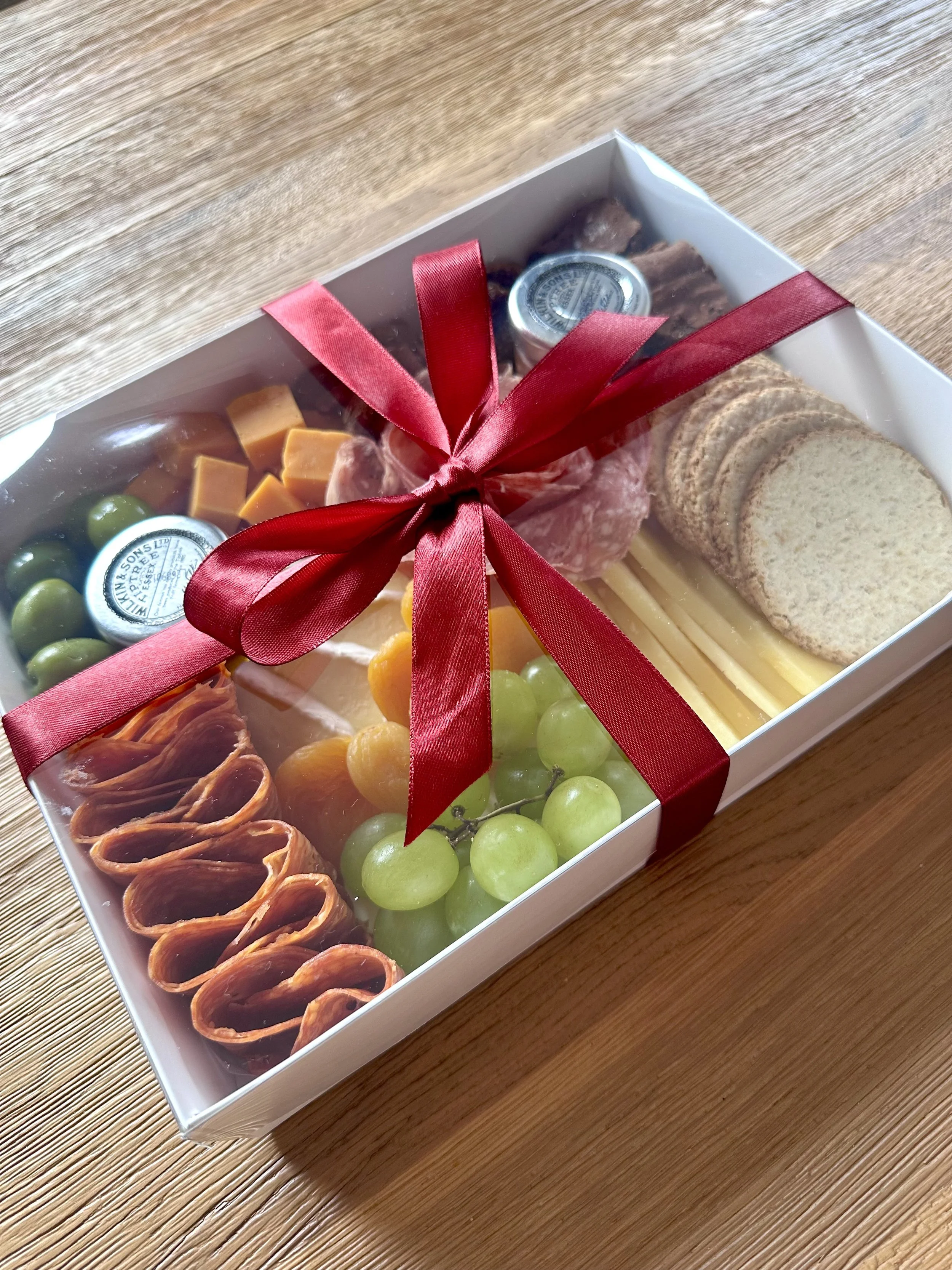 A gift box with a burgundy ribbon containing assorted cheeses, green grapes, oatcakes, sweet treats, and slices of deli meat, placed on a wooden surface.