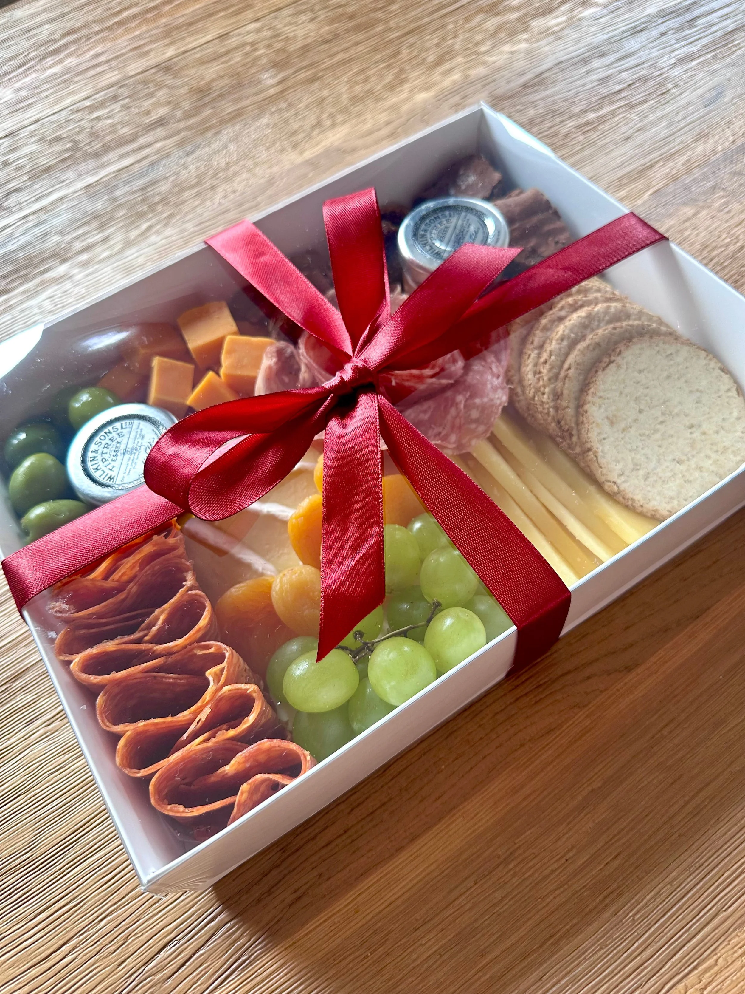 A gift box containing charcuterie and sweet treats, tied with a burgundy ribbon, placed on a wooden surface.