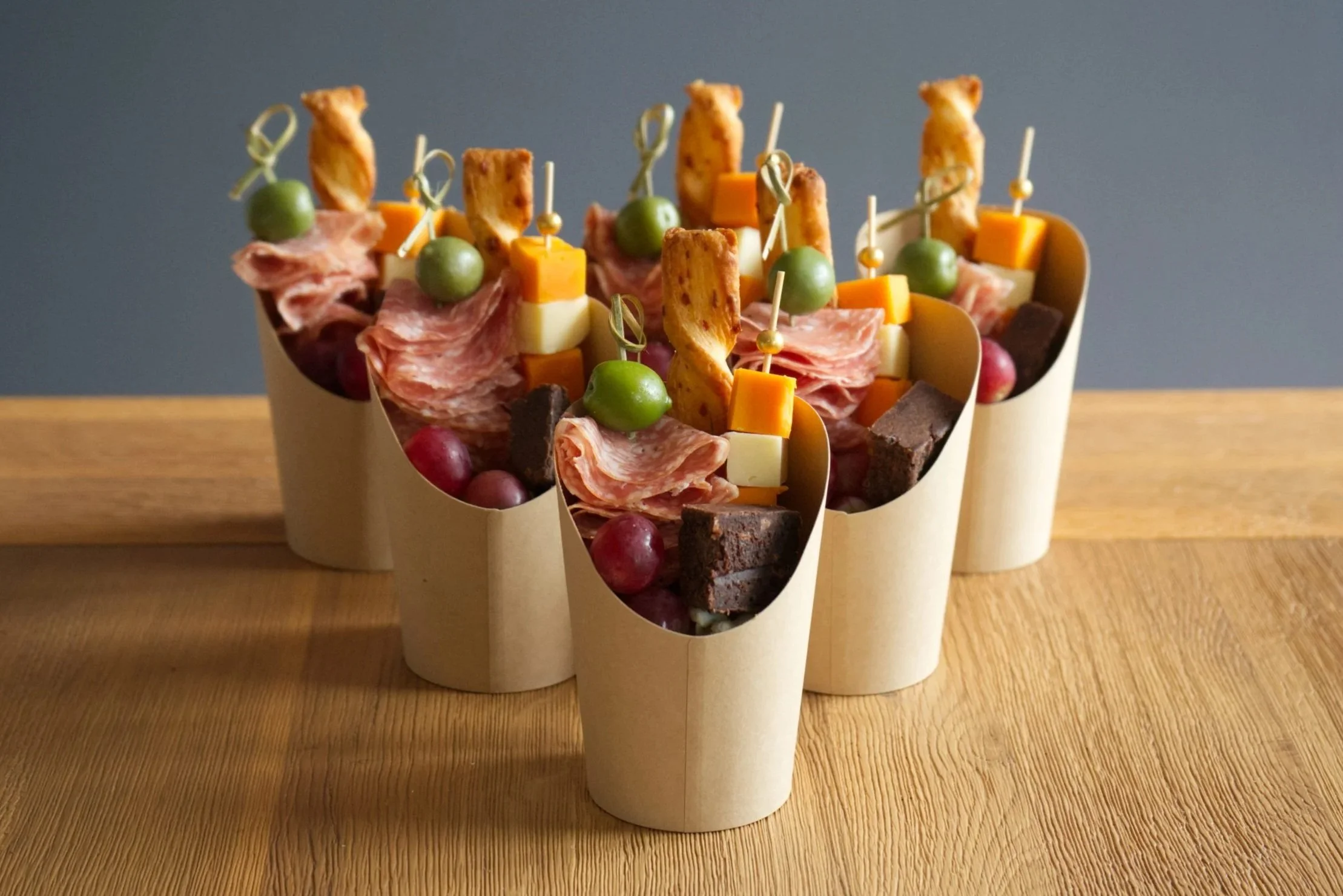 Close-up of several paper cups filled with assorted party snacks including cheese cubes, green olives, grapes, slices of cured meat, and pieces of chocolate brownie, arranged on a wooden surface against a gray background.