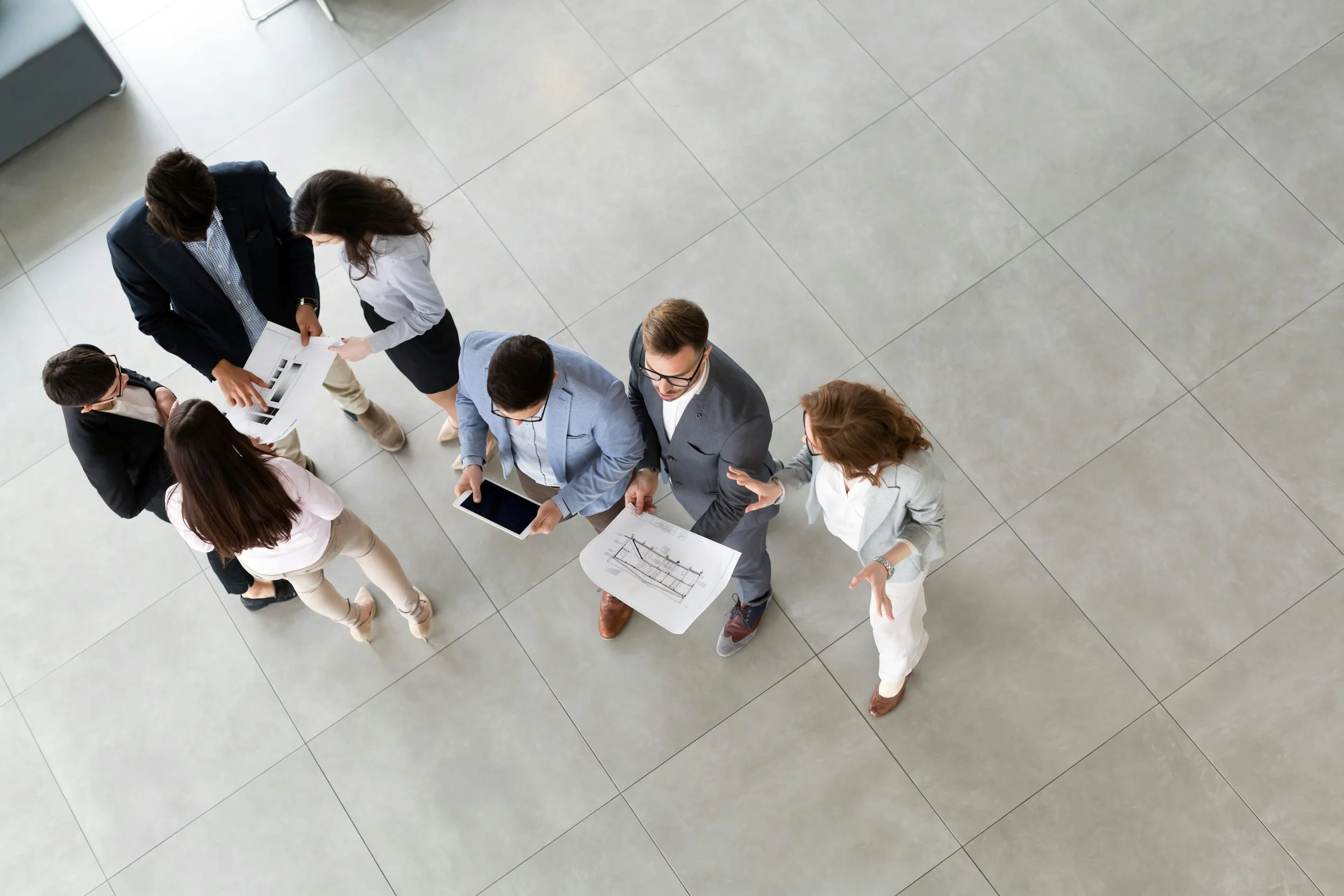 A group of professionals standing in a modern office lobby, looking at documents and discussing, viewed from above.