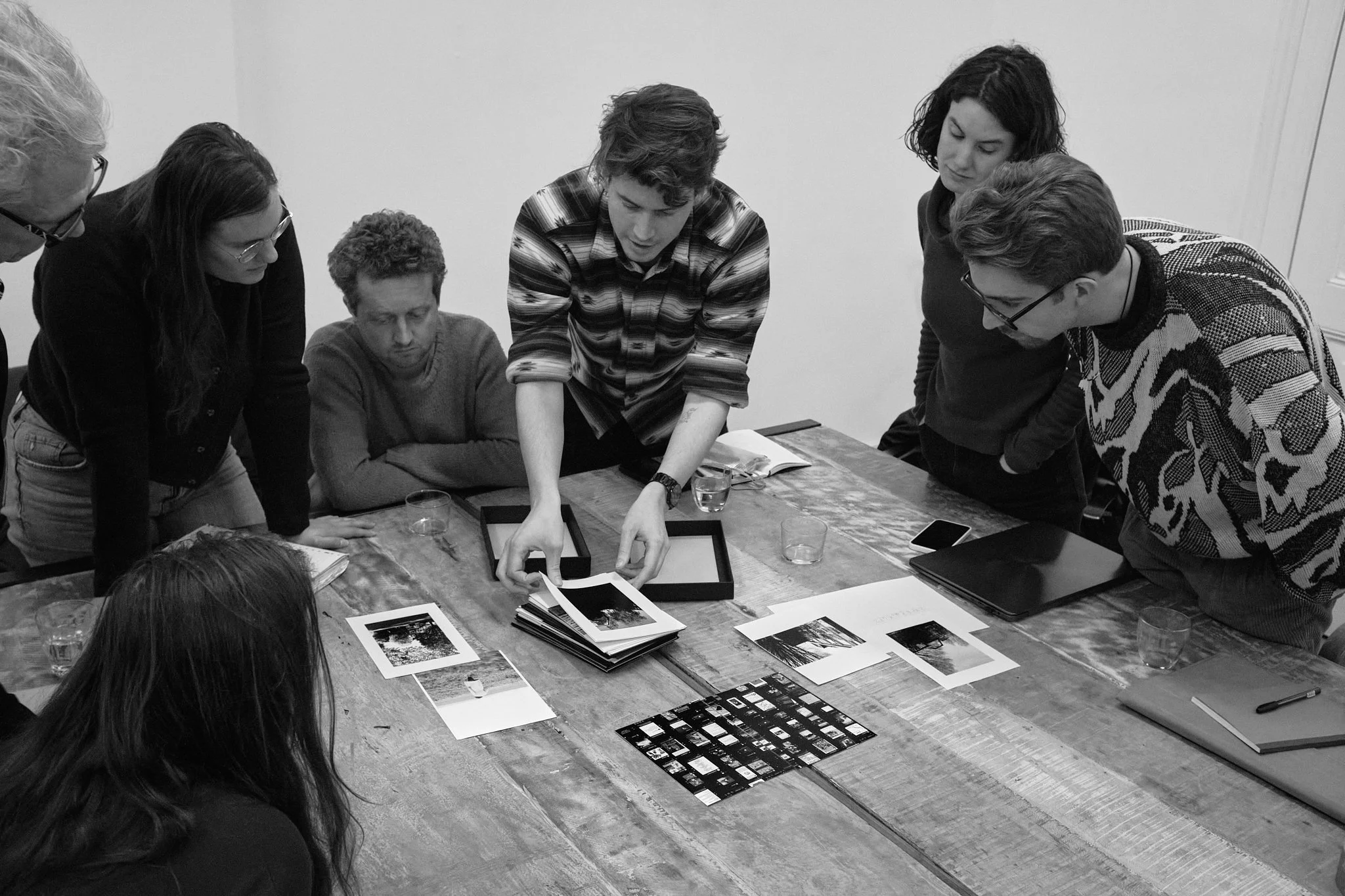A group of makers gathered around a table, looking closely as one participant opens a photobook and points to a print.