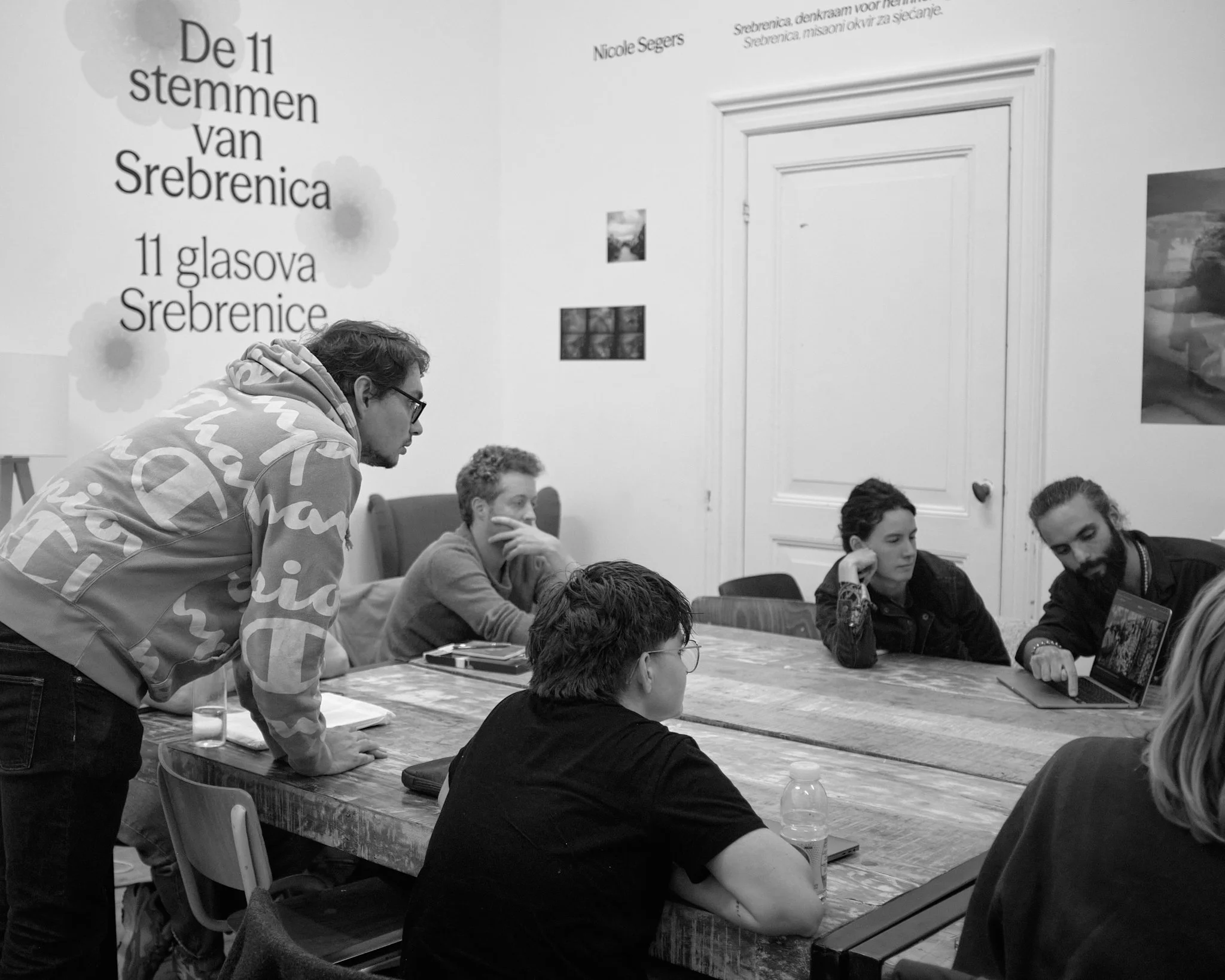 A Wanderstories session at Fotodok: a photographer leans on a large wooden table to speak with seated participants, an exhibition text on the wall behind them.