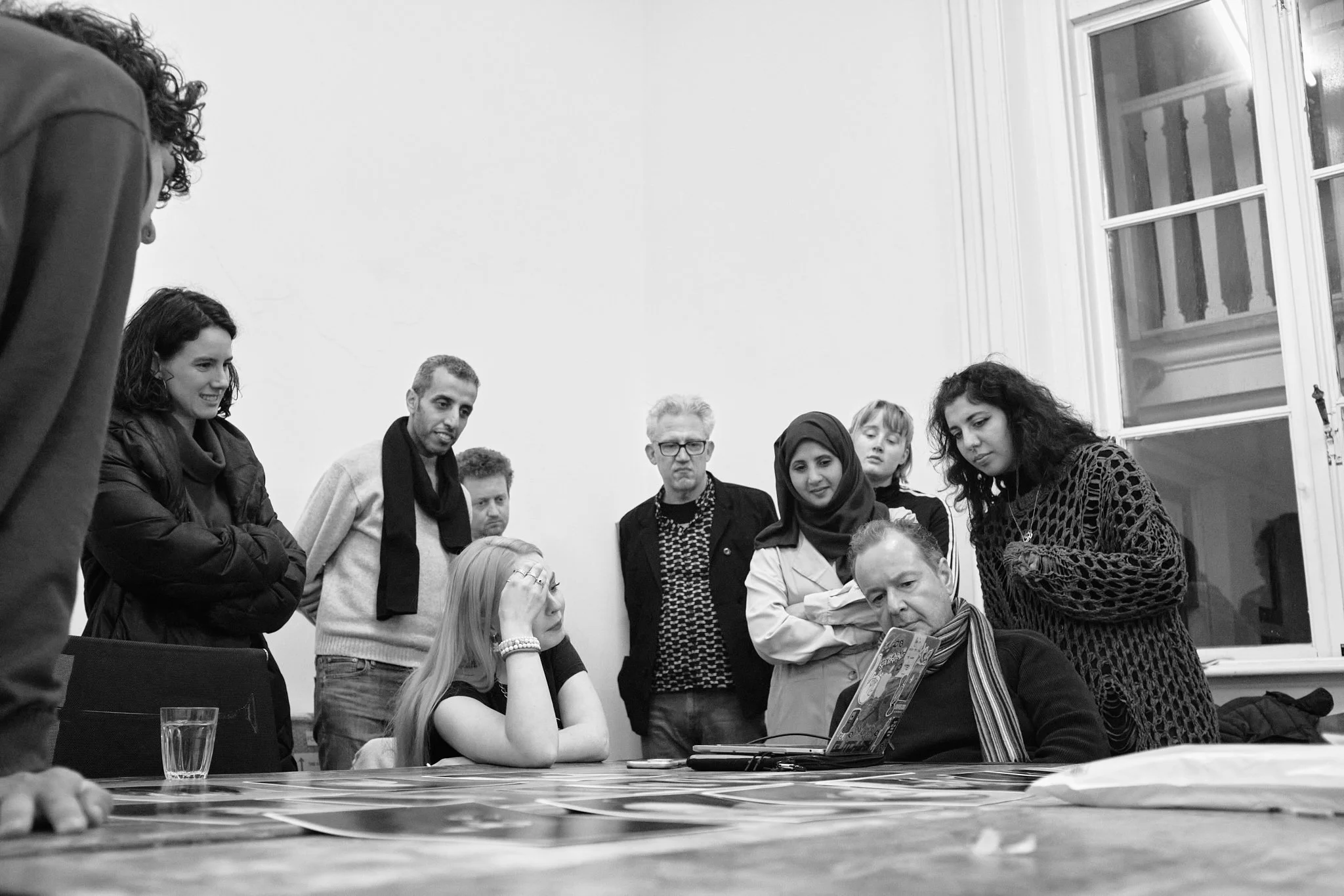 A wider view of a Wanderstories session: a circle of participants looks down at work on the table, lit by daylight from a tall window.