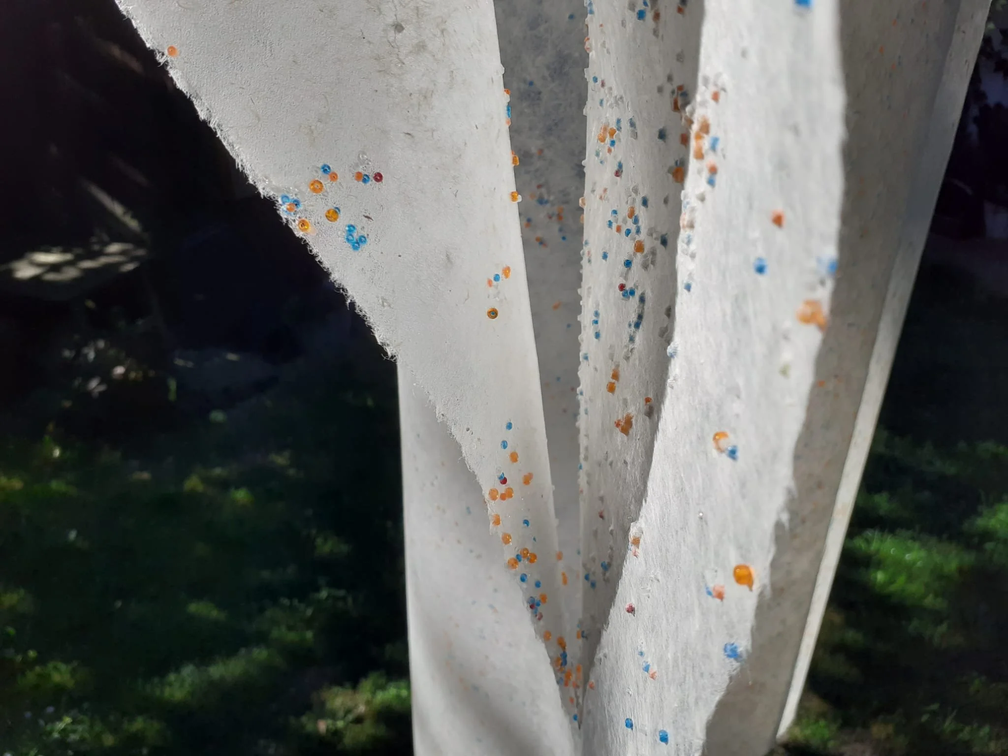 Close-up view of a white paper or cardboard surface with scattered colorful small circular confetti in blue, orange, and red. The surface appears to be torn or cut with uneven edges. The background is dark and slightly blurred, suggesting the surface is outdoors with grass or foliage nearby.