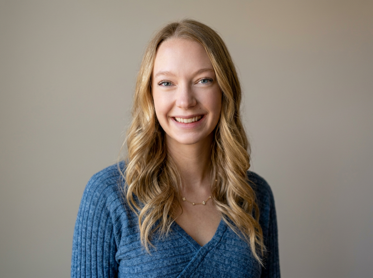 A young woman with long, wavy blonde hair, smiling at the camera, wearing a blue sweater and a delicate necklace, standing against a neutral beige background.