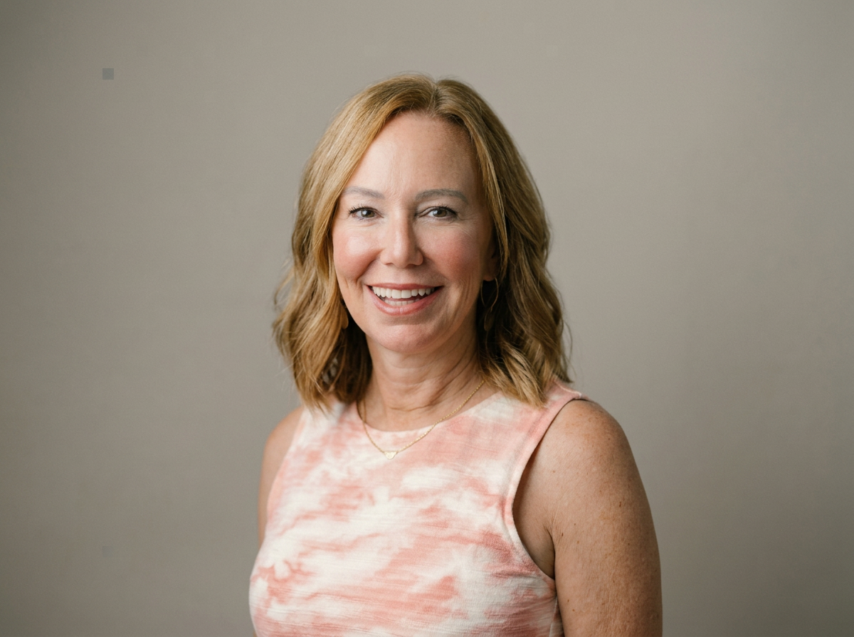 A woman with shoulder-length, wavy, blonde hair wearing a sleeveless, light pink, and cream tie-dye top and gold jewelry, smiling at the camera against a neutral background.