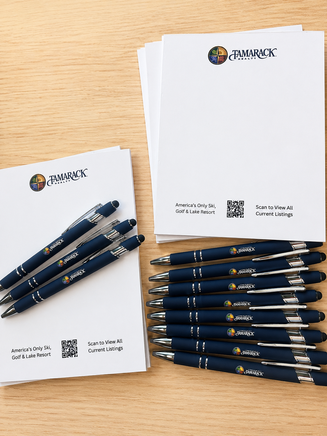 Promotional materials from Tamarack Realty, including branded pens, brochures, and notepads on a wooden desk, with text indicating 'America's Only Ski, Golf & Lake Resort' and a QR code to view listings.