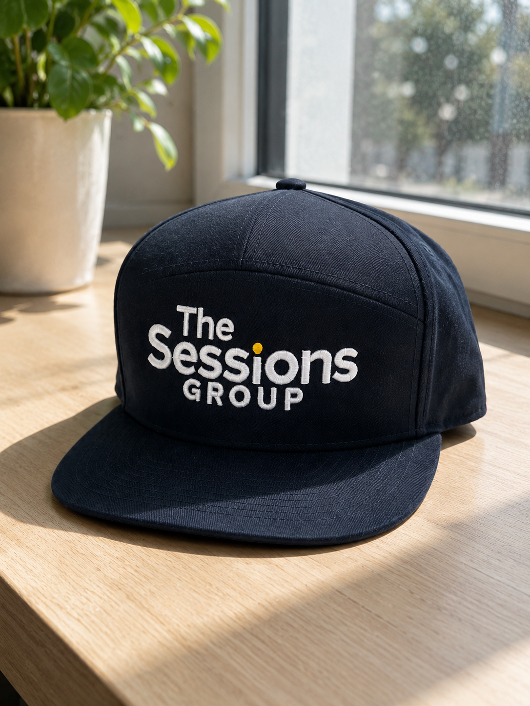 Black baseball cap with white embroidered text that reads 'The Sessions Group' placed on a wooden surface near a window with a plant in a pot nearby.