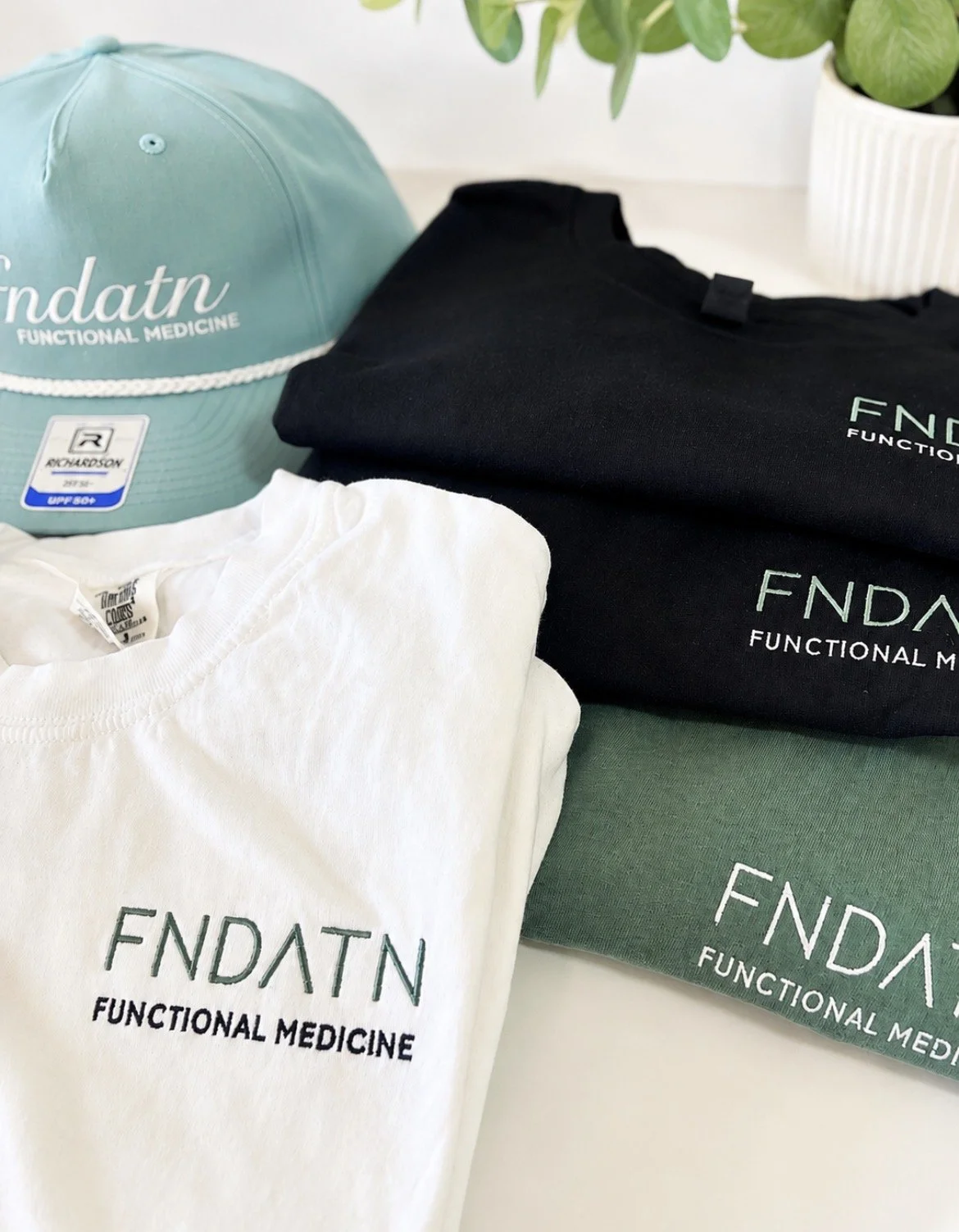 A light blue baseball cap with white text reading 'FNDATN FUNCTIONAL MEDICINE', white and black folded T-shirts with similar text, and a potted plant with green leaves in the background.