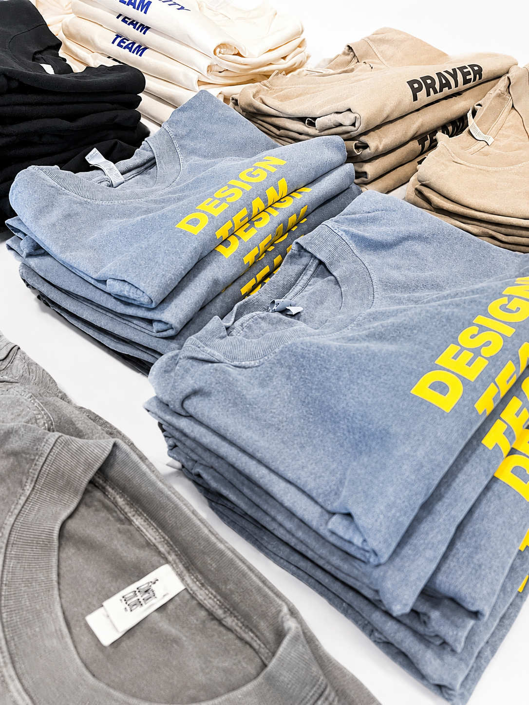 Stacks of folded hoodies and sweatshirts in various colors with printed text, including yellow "DESIGN TEAM" and black "PRAYER" on beige and blue garments, displayed on a white surface.