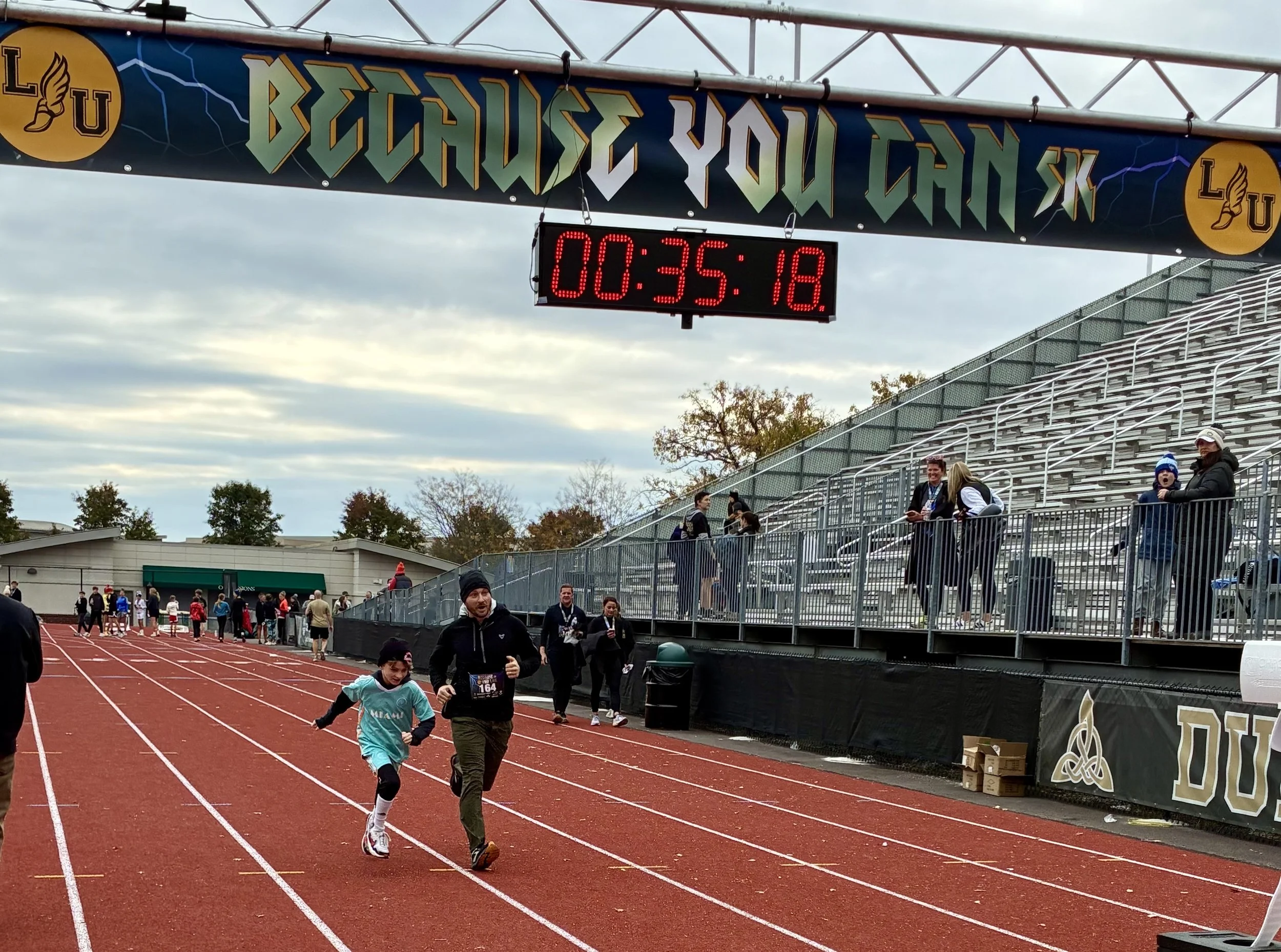 People running on a track at a stadium with an electronic timer display showing 00:35:18 and a banner above that reads 'VOORDEVOE CANDX'.