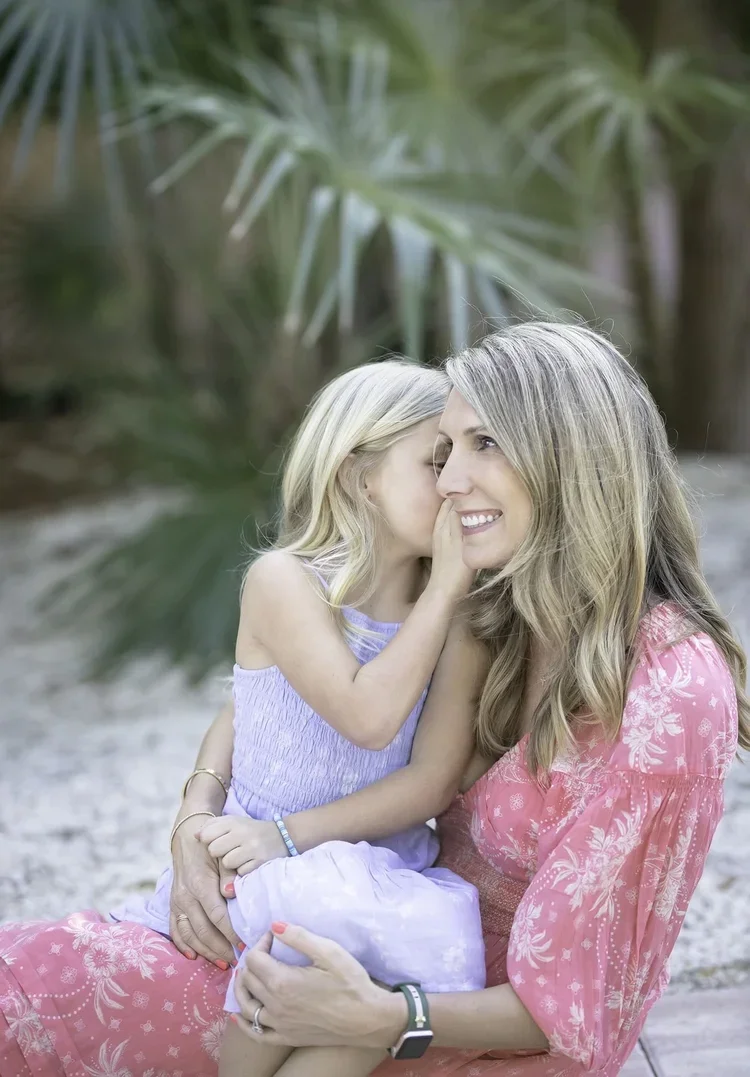 A young girl whispers into a woman's ear, who is smiling, while sitting outdoors with palm trees in the background.