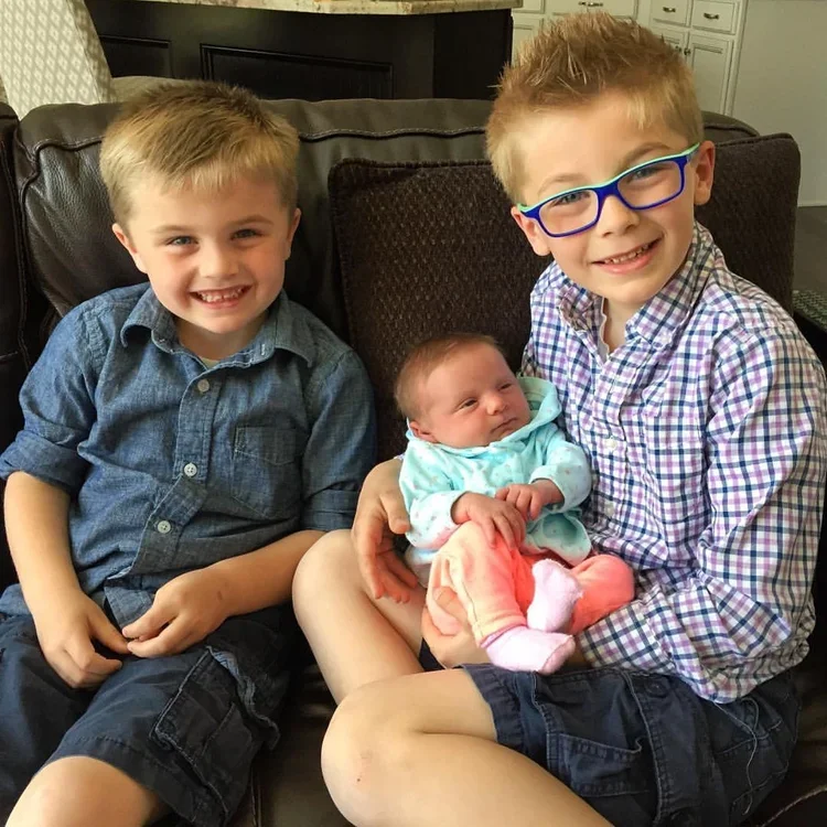 Two young boys, one wearing glasses, holding a newborn baby girl on a brown couch in a living room.