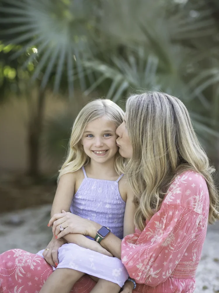 A woman with long blonde hair wearing a pink floral dress kisses a young girl, who has blonde hair and blue eyes, on the cheek while sitting outdoors with green foliage in the background.