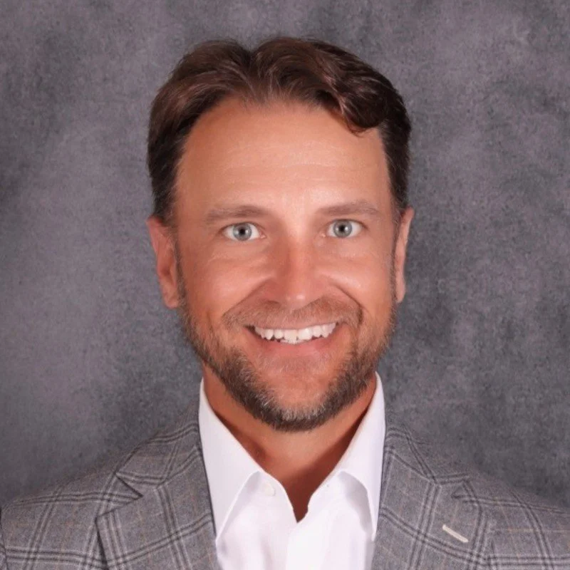 A man with brown hair and a beard, smiling, wearing a white shirt and a gray plaid blazer, against a gray background.