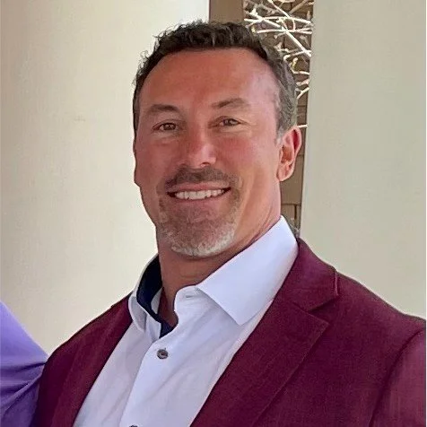 Smiling man with short dark hair, light facial hair, wearing a white collared shirt and burgundy blazer.